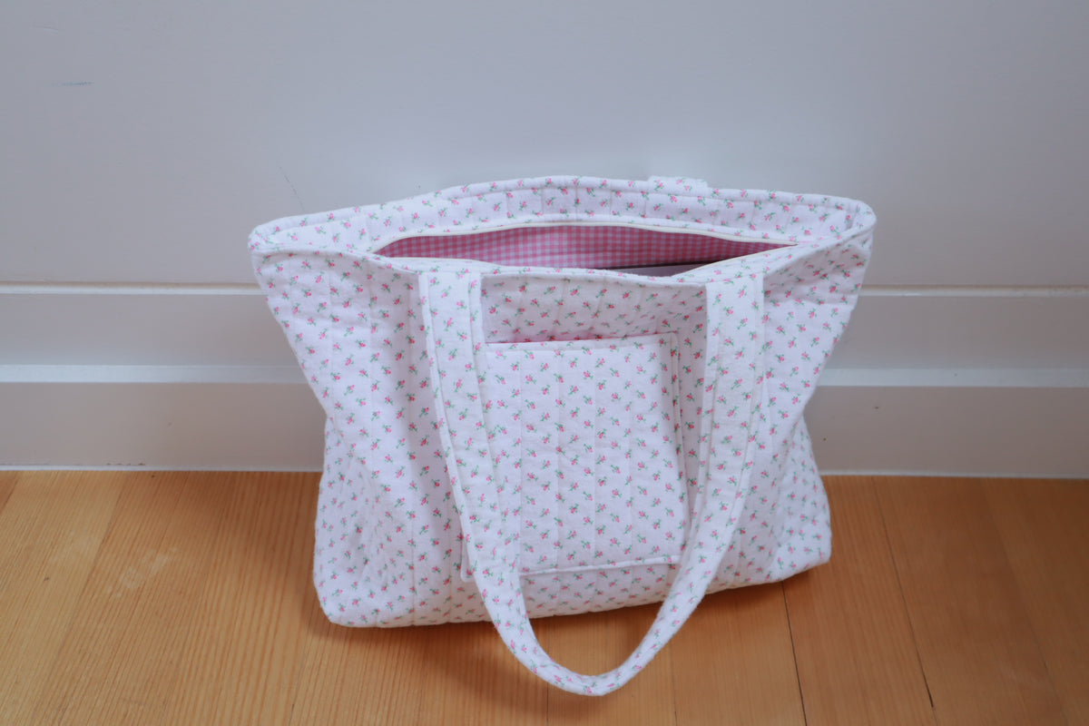 A white fabric tote bag with a small pink floral pattern, featuring two shoulder straps, a zipper, and a front pocket, sitting on a wooden floor against a white wall.